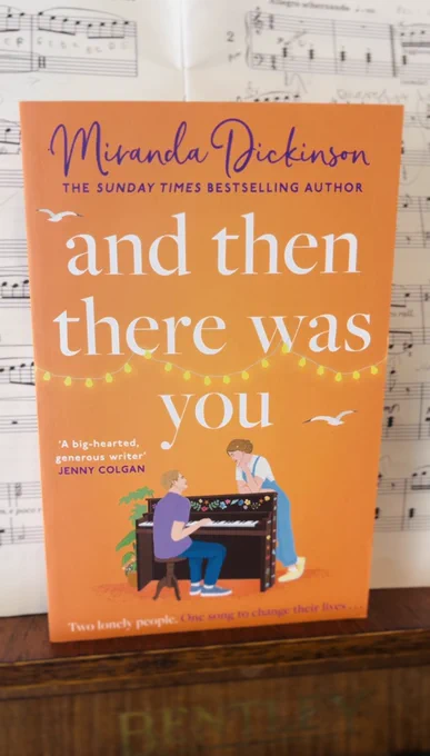 A paperback of my brand new book, And Then There Was You, resting on the music stand of my piano, with sheet music behind it. The cover shows a man sitting at a painted piano with a woman leaning against it, listening to him play. Strings of white lights loop above with two seagulls flying. The tagline reads: “Two lonely people. One song to change their lives.”