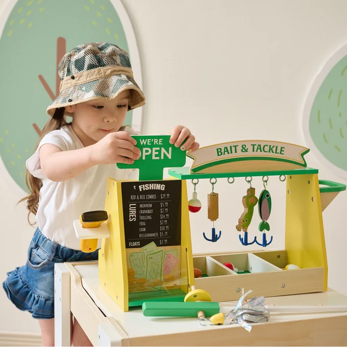 A child plays with a wooden "Bait & Tackle" fishing shop playset, featuring colorful fishing rods, fish, and a cash register. The setup includes a "We're Open" sign and is set against a room with a pastel-colored, circular pattern on the wall.