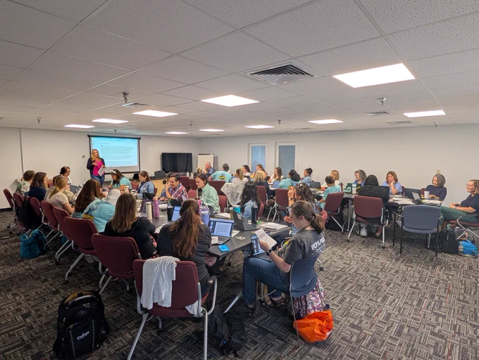 A large group of people are meeting. They are seated in two columns of tables with computers in front of them. A presenter is at the front of the room.