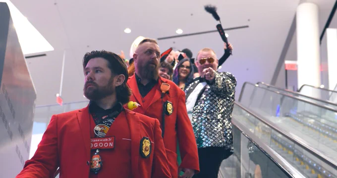Alt text: Lintile, wearing a silver sequin jacket and gold sunglasses, rides an escalator down with a “goon escort” to Hacker Jeopardy. Two men in matching red suits with badges and “GOON” name tags lead in front, looking serious. Lintile points toward the camera while holding a black prop, with other attendees in colorful attire smiling behind. The scene is brightly lit in a modern convention space.