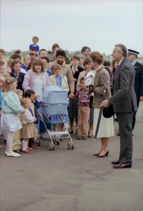 Princess Anne standing on the airfield tarmac with members of the public, including children, standing to her left ready to meet her.