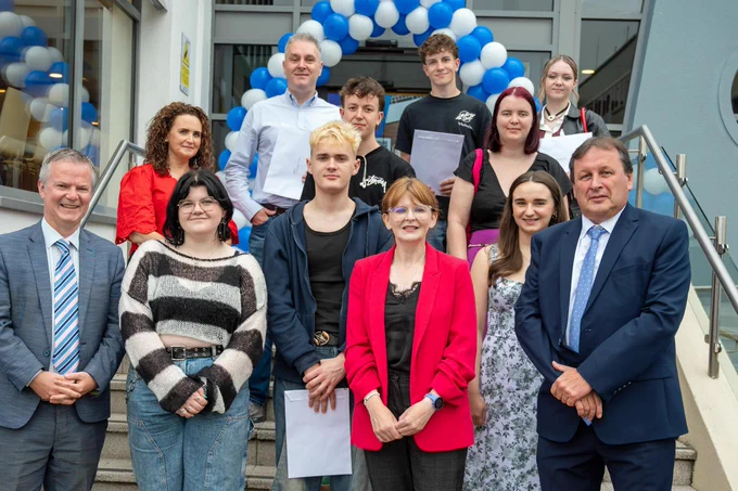 A photo of the Minister with a a group of people at the steps of the North West Regional College in Derry with a balloon arch in the background as young people get their exam results.