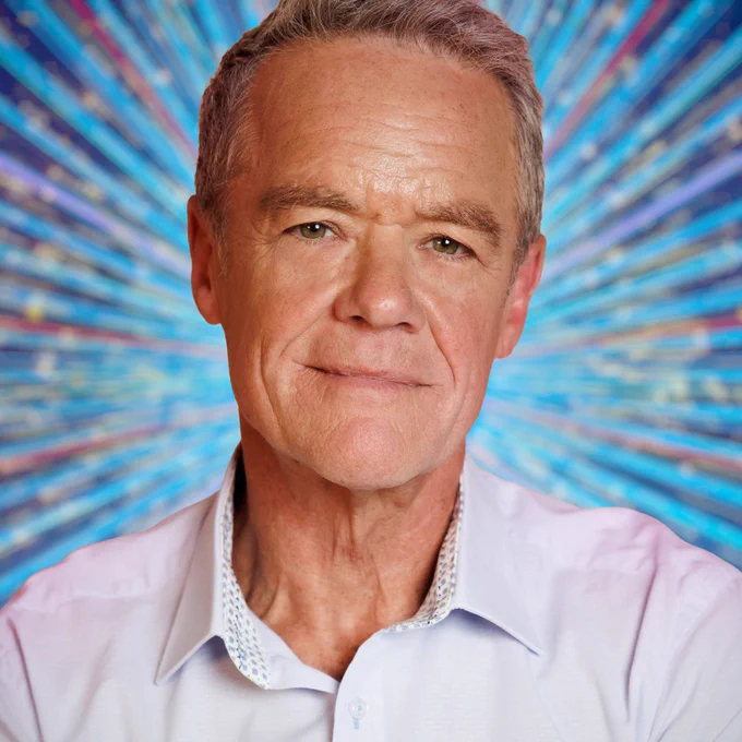 Stefan Dennis smiles to camera in a light shirt, set against a glittering blue background.