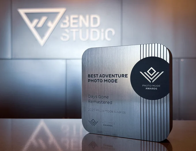 A small square trophy sits on a tabletop on the left side of the image. On the trophy it reads, “Best Adventure Photo Mode. Days Gone Remastered”. A large white Bend Studio logo can be seen in the background on a gray aluminum wall.