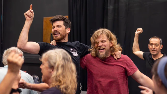 Exe Men rehearsal photo. The cast celebrate and chant together. One man has his arm around another’s shoulder while chanting and pointing upward.