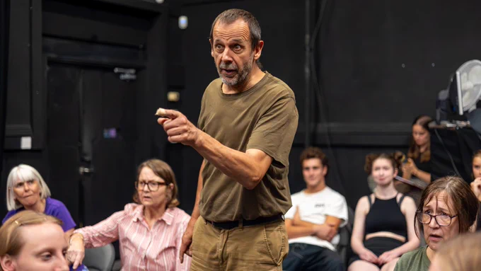 Exe Men rehearsal photo. Cast members sit in rows as one man stands, pointing forward with a serious expression.