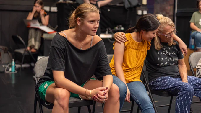 Exe Men rehearsal photo. Three cast members sit in a row. One girl stares intently ahead while the other two share a supportive hug.