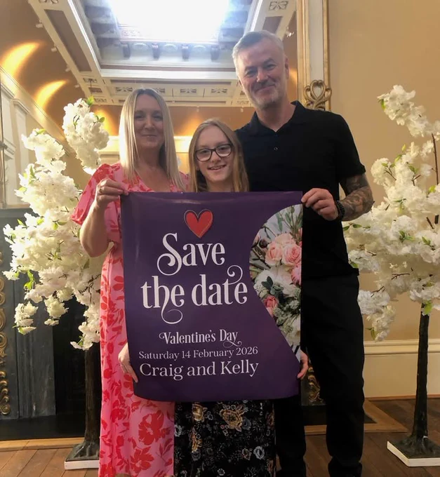 Kelly, Craig and their daughter holding a save the date sign inside drapers hall