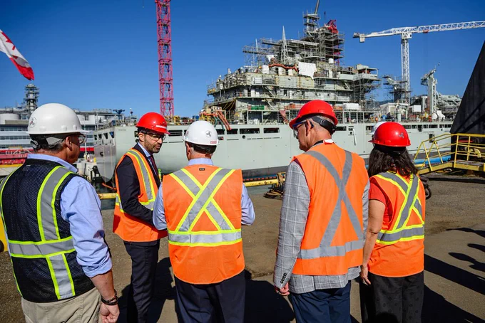 A man in a red hard hat is talking to a group of 4 other people also wearing safety vests and hard hats, in front of a large ship.
