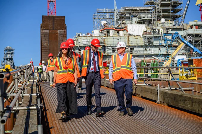 A group of people walk down a boat dock wearing safety vests and hard hats. A giant ship is behind them.