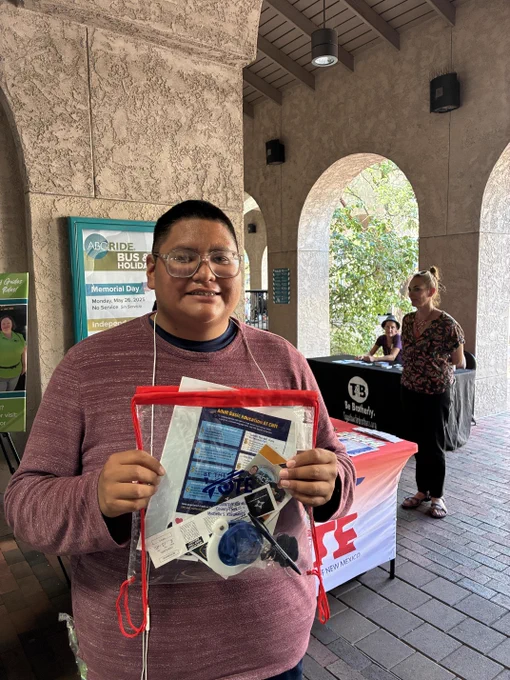 Person wearing a maroon sweater holds a clear red-trimmed pouch filled with voting and educational materials in a covered outdoor area with stone arches.