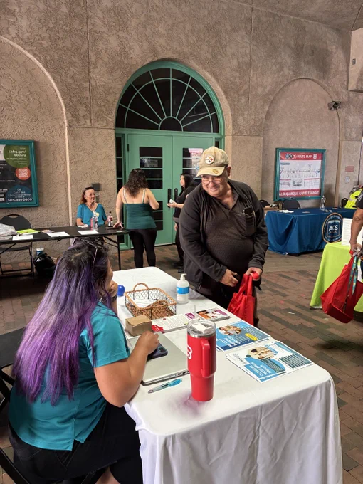 A woman with long purple hair sits at a table with informational pamphlets and a laptop, engaging with a man holding a red bag in an indoor event space.