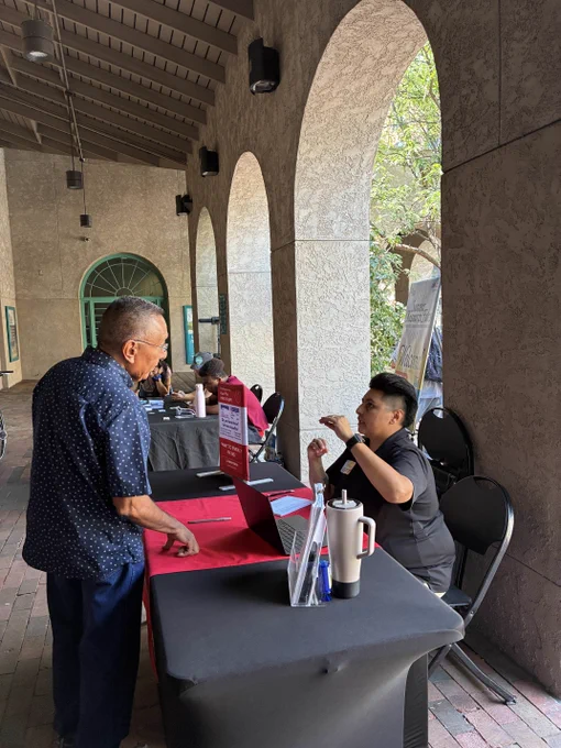 Man in a blue shirt talking to a seated woman at a registration table under stone arches with a laptop and informational signs.