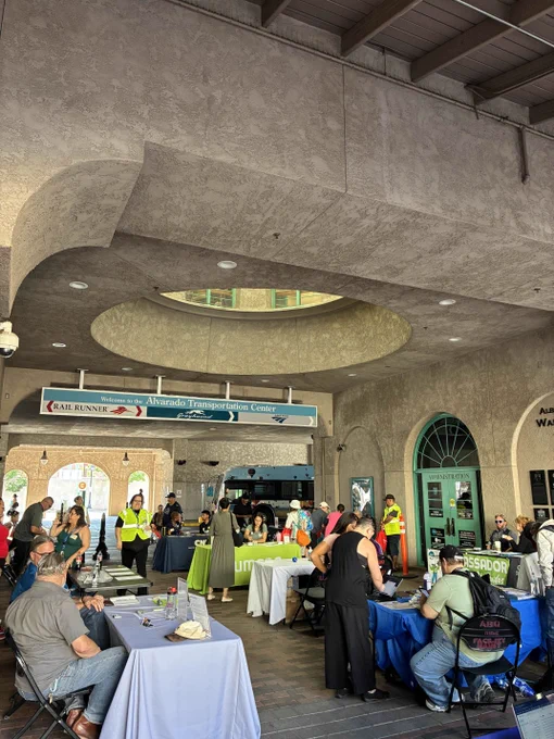 Busy scene inside Alvarado Transportation Center with multiple information tables and people interacting under a textured concrete ceiling.