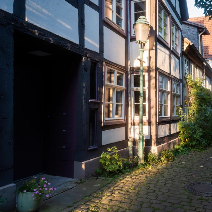 A captivating street scene unfolds, featuring a traditional European half-timbered building with a prominent, antique lamppost bathed in the warm, golden light of late afternoon. A rustic cobblestone path leads into the frame, adorned with patches of green grass and a small pot of flowers. The dramatic interplay of sun-drenched facades and deep, cool shadows creates striking contrast and depth, with a mysterious, dark doorway adding a touch of intrigue. — Warm light finds old post, Shadows deepen on the path, Time whispers softly.
