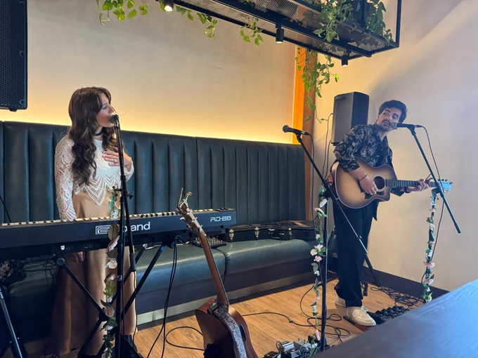 Two people performing on a makeshift stage. One is standing in front of a keyboard piano and singing into a microphone. The other is playing an acoustic guitar and singing into the mic while looking at the other person.
