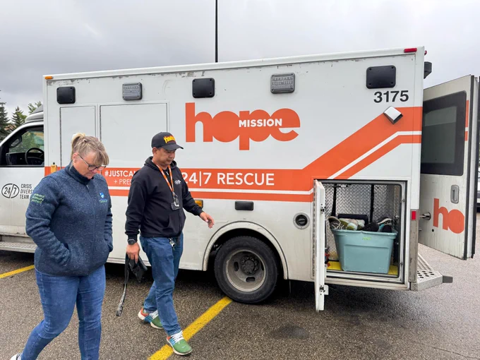 Two people walking next to a 24/7 rescue vehicle that says "hope mission; 24|7 rescue) in orange. Part of the vehicle is open.
