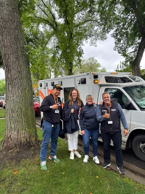 A group of four people smiling and standing in front of a crisis response vehicle and a large tree.
