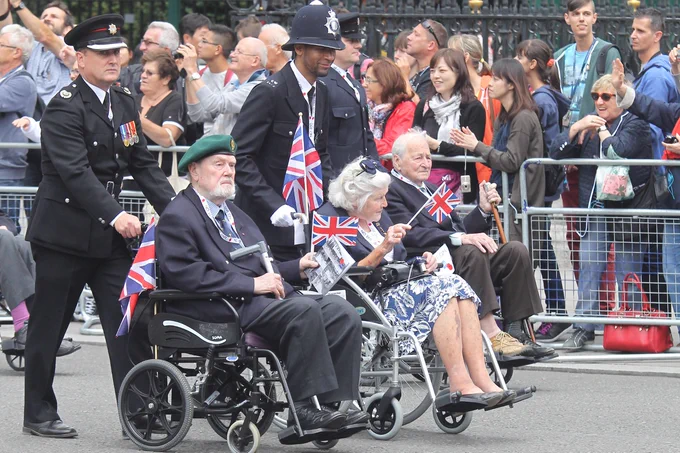 A group of three veterans in wheelchairs being pushed by Metropolitan Police officers during a parade to Westminster Abbey