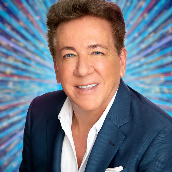 A head and shoulders shot of Ross King, who wears a navy suit and white shirt, set against a glittering blue background