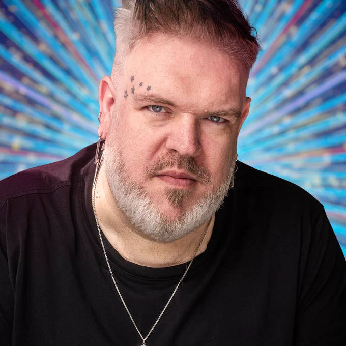 A head and shoulders shot of Kristian Nairn set against a glittering blue background