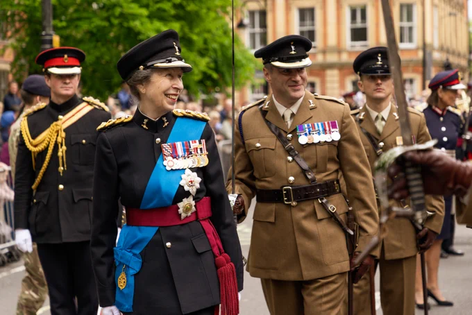 Princess Anne shown in her ceremonial uniform, with a medal rack on her chest, smiling to soldiers not photographed. There are soldiers in brown uniforms following behind her.