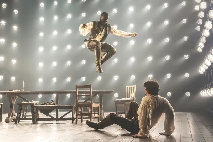 Ncuti Gatwa and Edward Bleumel in Born With Teeth. They wear period clothing, Ncuti in all black leathers as he jumps high off a wooden table toward Edward, sat scared on the floor with his back to the audience. The entire background is spotlights.