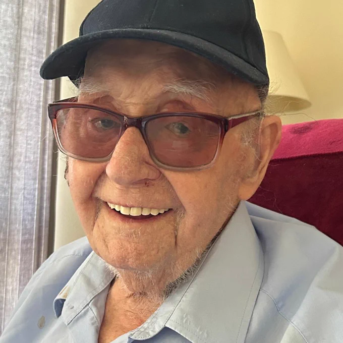 An image of Tom, a 103 year-old Second World War veteran, sitting in a chair, smiling at the camera wearing a light-blue shirt, red-tinted glasses and a black cap.