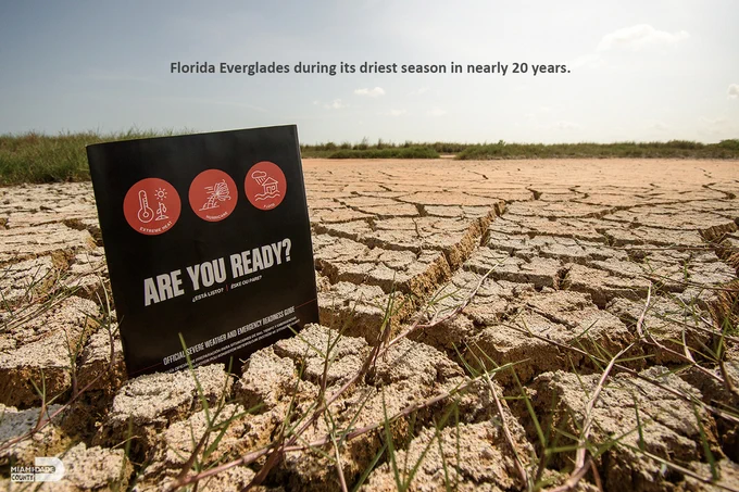 The ground which is extremely dry in the Everglades. It is an orange color with cracks all through it.  There is an Extreme Weather Guide sticking out of it that is black with red circles and white writing. 