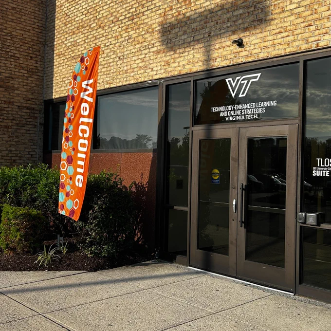 The entrance to the TLOS office suite 21 at University Mall. There is a VT logo with Technology-enhanced Learning and Online Strategies, Virginia Tech above the double glass doors and an orange feather shaped flat with the word Welcome and a maroon, teal, and white hexagon pattern.
