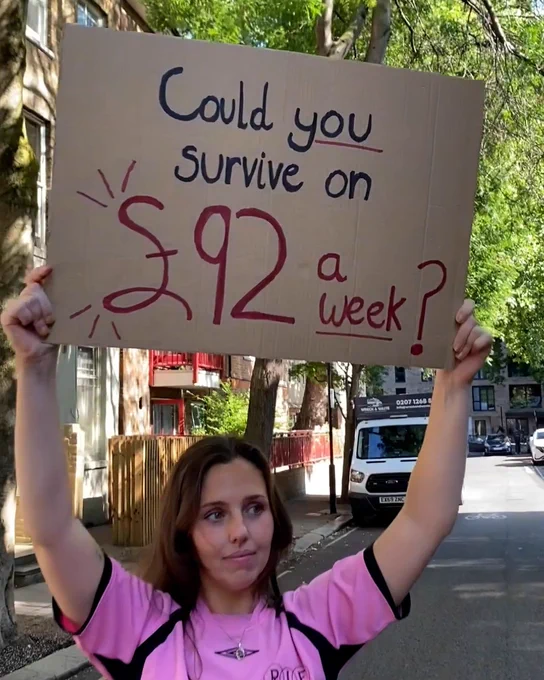 Photo of a white woman with brown hair, wearing a pink t-shirt. She stands in a street in London holding up a hand-written, cardboard sign which reads: 'Could you survive on £92 a week?'