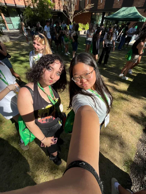 Two individuals wearing conference lanyards taking a selfie in a sunny outdoor setting with other attendees in the background.