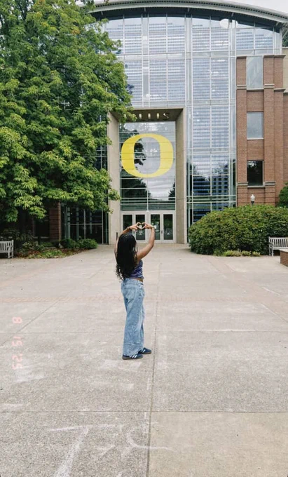 A person stands in front of the University of Oregon's Lillis Business Complex, capturing a photo of the building's large glass facade with an "O" logo.