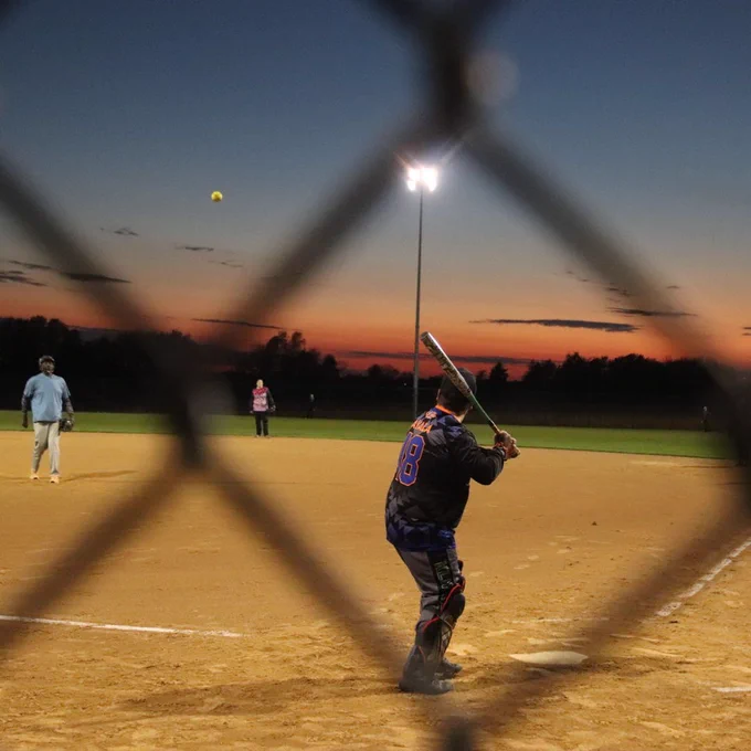 Looking through a chain link fence a batter gets ready to swing the bat as the ball comes his way. The sky is lit with color as the sun sets.