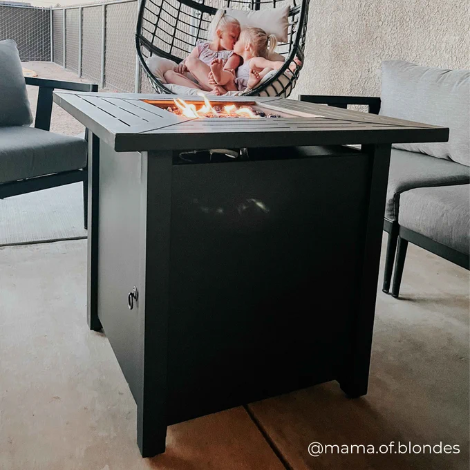 Fire pit table with visible flames, located on a patio with a seated adult and child in the background, enjoying the warmth.