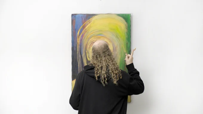 Archie Barry, Dream for Reed (2025), video still. Courtesy of the artist.
[Archie Barry wears a black hoodie and stands facing a colorful painting with swirling yellow, green, and purple tones. They point towards the artwork with one hand, against a plain white wall background.]