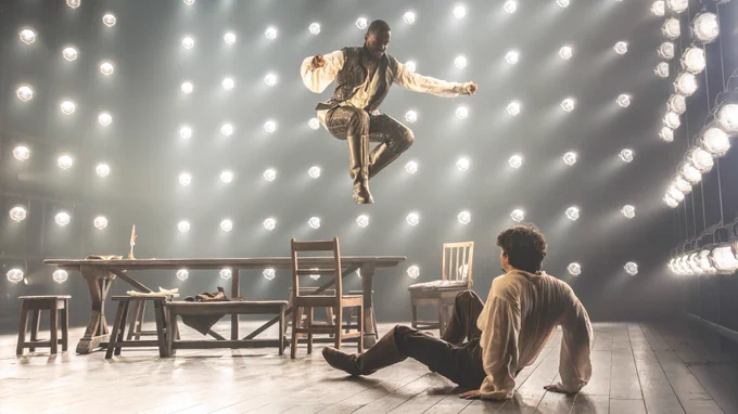Ncuti Gatwa leaping above a wooden table and chairs. Edward Bluemel is sat on the floor, leaning back
