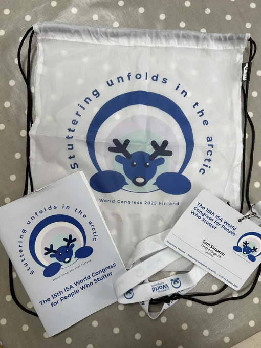 Photograph of the 15th ISA World Congress for people who stutter programme positioned on top of the conference drawstring bag adjacent to my conference lanyard. All three are white & feature a marine blue reindeer head around which the words ‘Stuttering unfolds in the artic’ are written