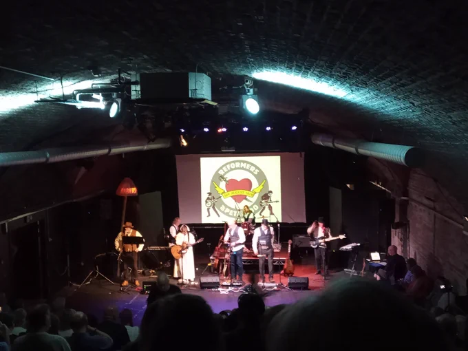Musicians in 19th-century costume on stage with guitars, keyboards etc, in front of the image of a heart pierced by a sabre, labelled "Reformers of Peterloo"