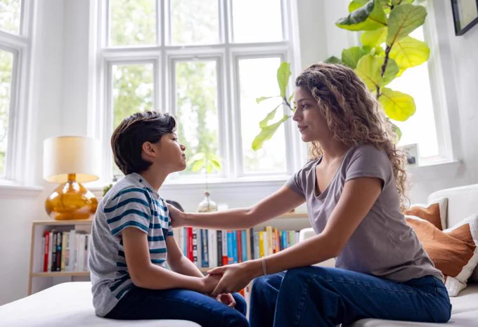 A person and a child are sitting on a bed, holding hands and looking at each other in a room with bookshelves and large windows.
