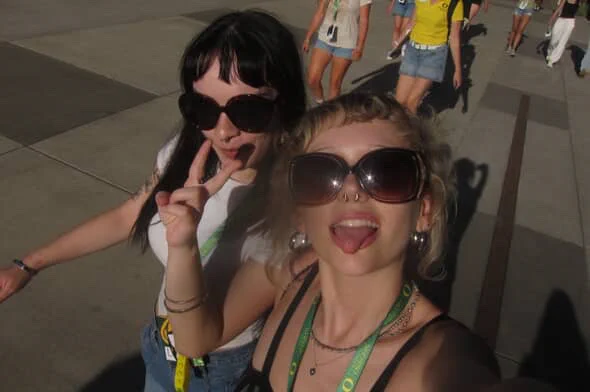 Two individuals in sunglasses making peace signs, enjoying sunlight, possibly at an outdoor event.