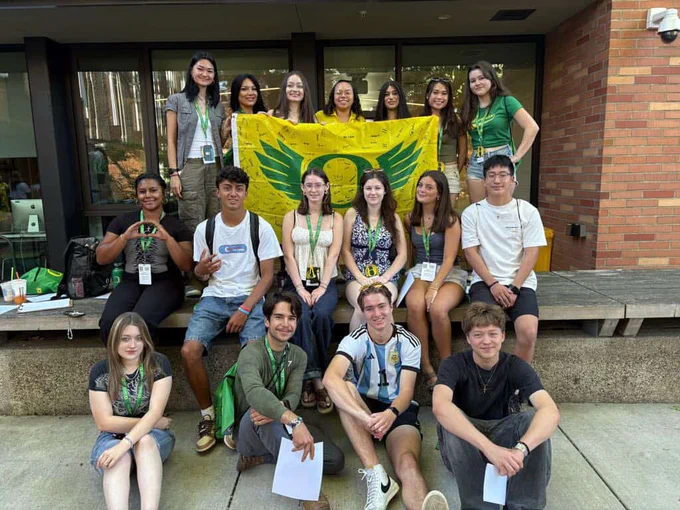 Group of students posing with a large Oregon Ducks flag in front of a building.