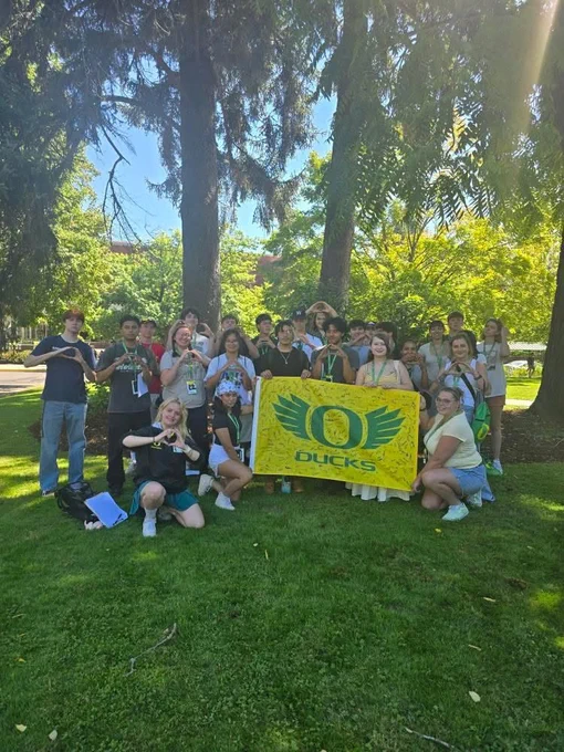 Group of individuals posing with a 'Ducks' banner on a sunny day on campus.