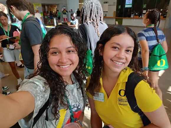 Two individuals, one wearing a camouflage top and the other in a yellow Oregon shirt, smiling at the camera in a crowded indoor setting.