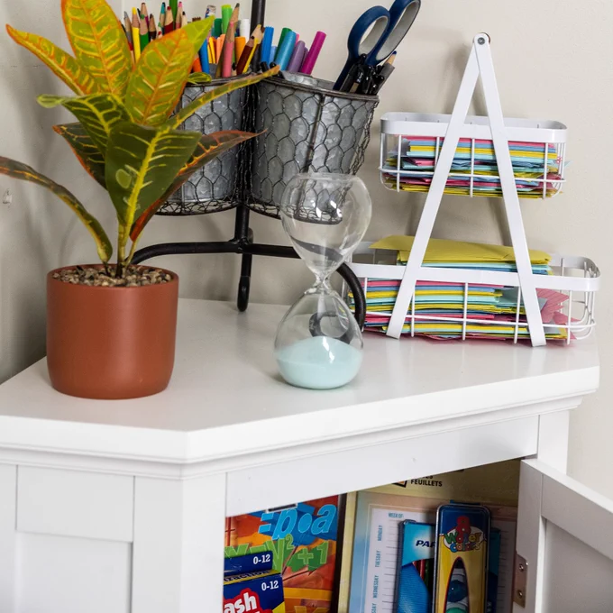 A neatly organized study area with a white desk containing a terracotta pot with a plant, a silver pot with a colorful foliage plant, and an hourglass. On a shelf above the desk are various stationery items neatly arranged, and below the desk are shelves stocked with magazines and books.