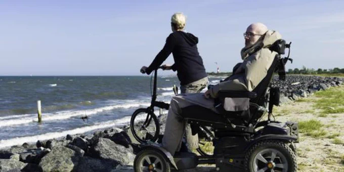 Two people enjoying a sunny day by the sea, one riding a bicycle and the other using a motorized wheelchair.