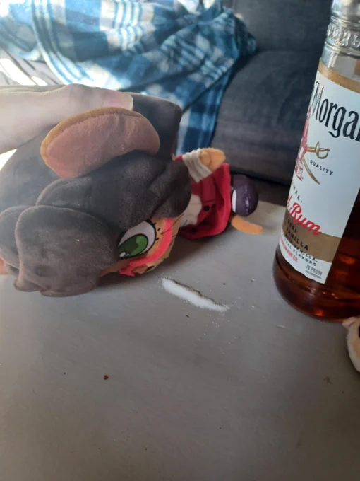 A plush of Margaret de Campos, a brown mouse girl in red glasses, lying face first on a table next to a line of white powder. A bottle or rum is next to her.