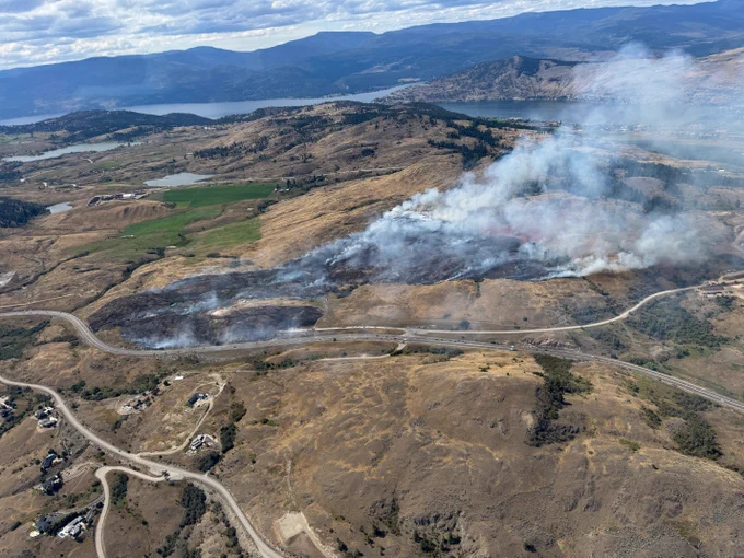 White smoke billows above a wildfire burning in the grasslands along a highway with a lake and rolling hills in the background