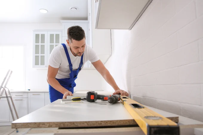 Person in work overalls measuring a piece of wood on a counter, with various tools around, in a bright kitchen setting.