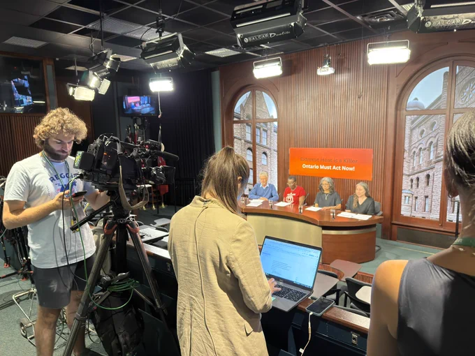 Photo from press conference inside the Ontario Legislature. Media are filming and writing in the foreground while four people sit at a presentation table with an orange sign behind them that says ‘Extreme Heat is a Killer, Ontario Must Act Now!’